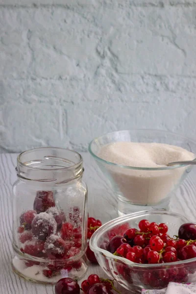 Currants and cherries in a glass jar, a glass plate with berries and sugar on the background of a table of white boards; light background for summer recipes for making homemade jam