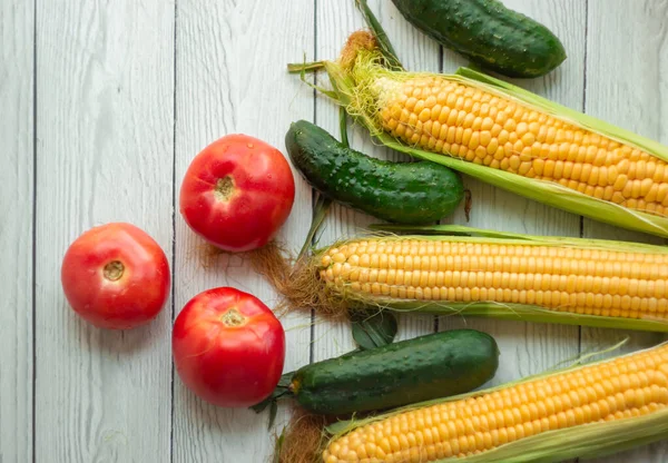 Ripe ears of corn, red tomatoes and fresh cucumbers lie on a light wooden table; vegetable bouquet, harvest