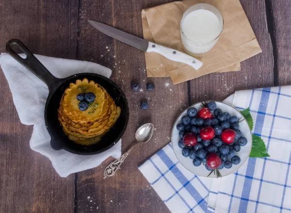 Pancakes with berries blueberries and cherries.pancakes in a frying pan on a wooden table with a plate of fresh juicy berries on a napkin,a glass of milk.Concept prepare a delicious Breakfast at home