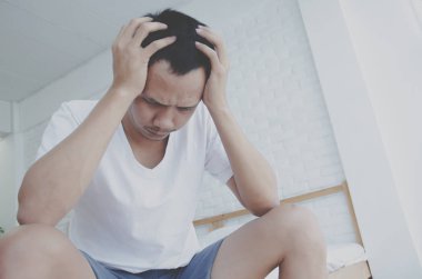 Erkekler odada yatağın kenarında oturuyor ve stres var. Beyaz gömlek giyen ve iki eliyle kafasında genç bir adam. Acı yüzünden..