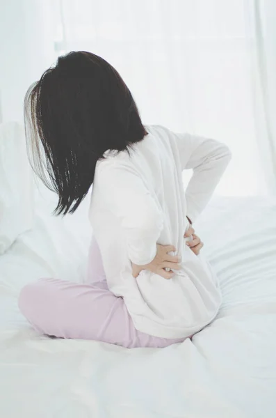 女性は朝 腰の痛みを伴う部屋でベッドの上で白いシャツとピンクのズボンを着用します アジアの女性は 朝に背中の痛みを持っています 背中の痛みはマットレスによって引き起こされる オブジェクトに焦点を当てない — ストック写真
