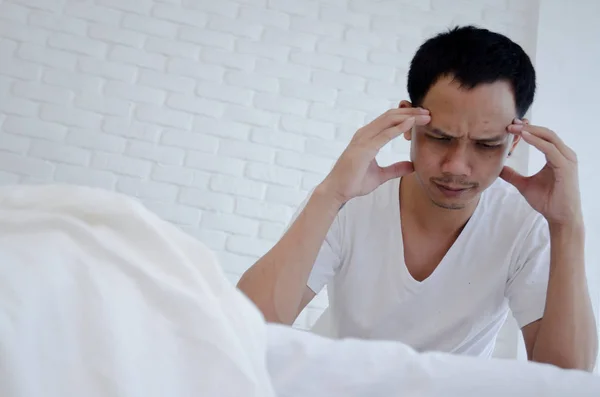 Men are sitting at the edge of the bed in the room and have stress.Young man wearing a white shirt with both hands on his head. Because of pain.