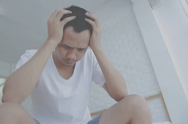 Men are sitting at the edge of the bed in the room and have stress.Young man wearing a white shirt with both hands on his head. Because of pain.