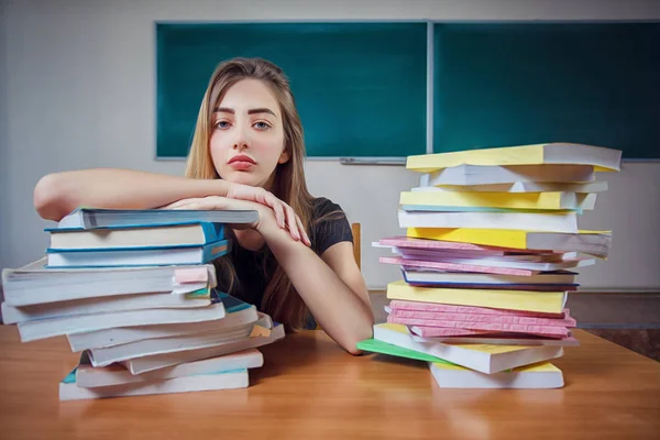Frustrated female student sitting at the desk with a huge pile of study books in classroom. Young college student at hard exam preparation in study hall looking tired and weary. Copy space
