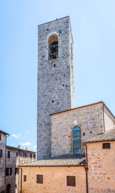 Dikey fotoğraf yüksek taş kule ile. Kule eski Unesco San Gimignano yeridir. Çan Kulesi üstünde var. Çevresinde çeşitli binalar vardır. Gökyüzü mavi ve açık.