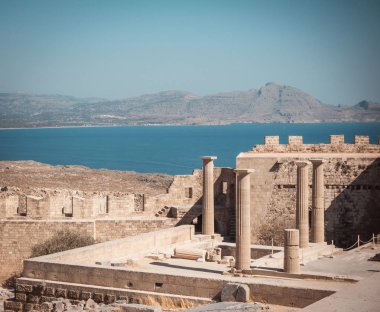 Lindos Acropolis birkaç ayağı ile yatay fotoğraf. Acropolys Akdeniz Rodos Adası üzerindedir. Deniz ve Gökyüzü mavidir. Birkaç kaya ve tepelerin arka planda görülebilir.