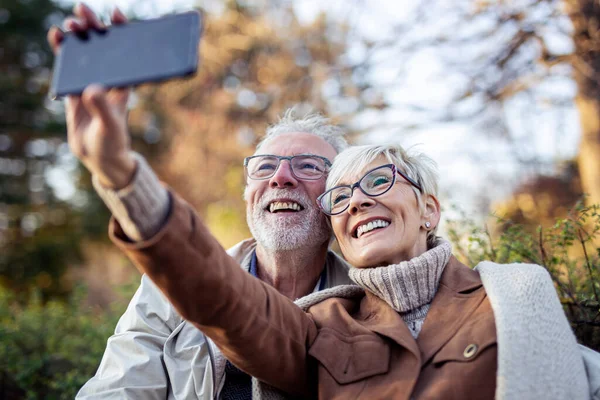 Yetişkin bir çift parktaki bankta otururken cep telefonuyla selfie çekiyor.