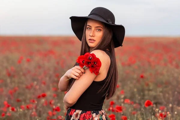 Beautiful long hair woman in black with hat on head at the middle of field full of red flowers