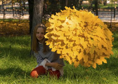 Güzel genç kız sonbahar sarı yapraklarından bir şemsiyeyle parkta yürüyor. Sonbahar zamanı, Eylül, Ekim, Kasım. Yapraklardan el işi. Şehirde yapraklar dökülüyor..