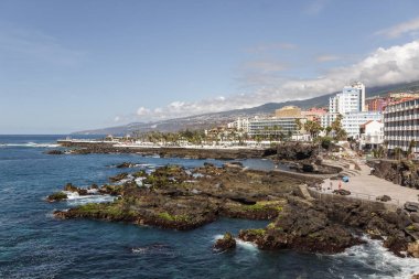 Lava rocks and cliffs along coast line at Puerto de la Cruz. Lagos Martianes in the background. Blue sky and beautiful clouds above mountains and hills of La Orotava valley. Tenerife, Spain clipart
