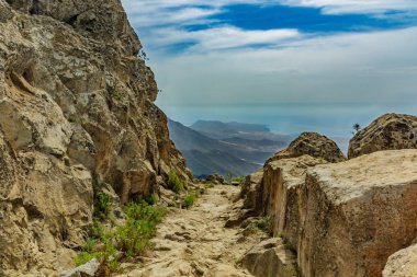 Güneşli bir günde çam ağaçlarıyla çevrili yayladaki taşlı yol. Berrak lue gökyüzü ve ufuk çizgisi boyunca bazı bulutlar. İğne yaprağı ağaçlarıile kuru dağ alanında Kayalık izleme yolu. Kanarya Adaları seyahat