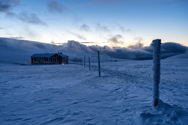 Mountain cabin with warm light in windows glowing in the dusk surrounded by snow landscape.