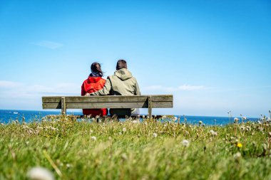 İki genç yetişkin, ahşap bir bankta deniz kenarında oturuyor ve güzel manzaranın tadını çıkarıyorlar..
