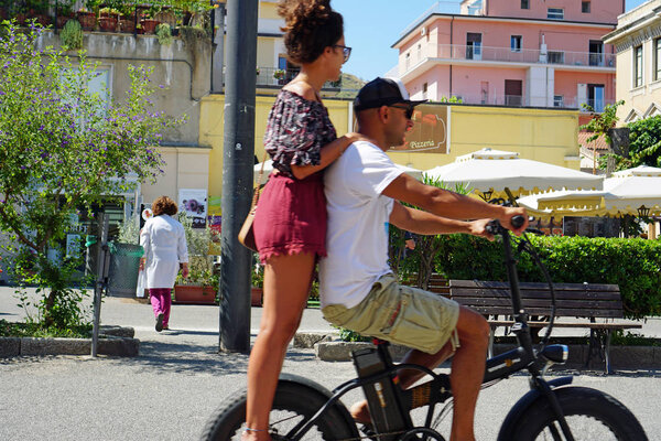 A young couple riding on one e-bike together, the girl standing on the back behind the rider