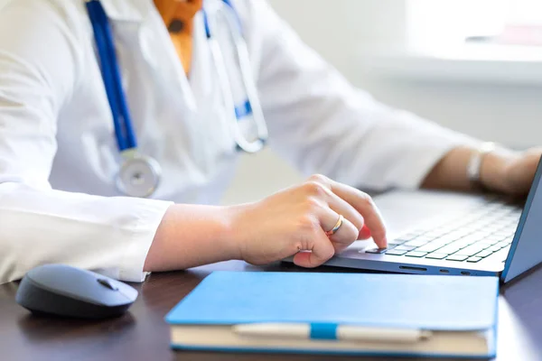 Beyaz laboratuvar önlüklü kadın doktor iş yerindeki masada dizüstü bilgisayarında not defteri ve tıbbi steteskopla yazı yazıyor. Tıbbi teknoloji, Elektronik Sağlık Kayıt Sistemi (EMR) kavramı. Uzaktan kumandalı bir doktor.