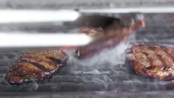 チーフは肉をトングでグリルに回す ジューシーで脂っこい揚げ肉 牛肉は大火で揚げられます フレーム内の火と煙の多く クローズアップスローモーション — ストック動画