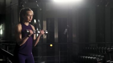 Genç ve güzel bir kız spor yapıyor. Dambıllarla hem sağ hem de sol elin iniş çıkışları.