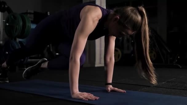穿着紫色紧身裤和T恤的漂亮年轻女孩在健身房里积极健身 双手放在地板上 把膝盖拉到肚子上 相机移动平稳 — 图库视频影像