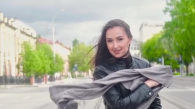 siyah deri ceket ince çekici kız Güneşli bir yaz gününde şehrin sokak boyunca yürüyor. O elinde büyük bir gri eşarp tutar ve kameraya coquettishly gülümsüyor. Güçlü rüzgarda çırpınan saçlar