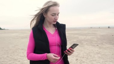 pembe t-shirt ve siyah yelek genç çekici kadın eğitim için telefonda müzik seçer. çalma listesi arasında kaydırma, iyi bir parça bulur. gülümsüyor ve uzağa bakıyor. ıssız kumlu plajda bulutlu yaz günü
