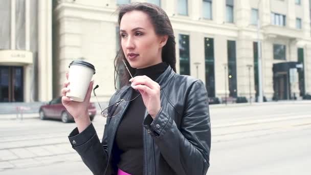 도시의 거리의 배경에 선글라스에 니블링 재킷에 아름다운 컵에서 커피를 마시다 — 비디오