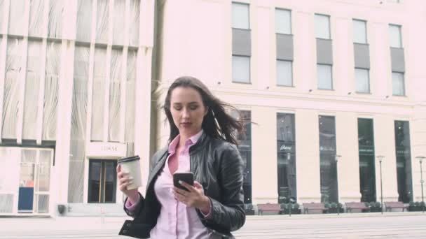 迷人的商务女孩站在街头背景 看着电话 喝着纸杯的咖啡 大风使她的头发发皱 背景中的城市街道 — 图库视频影像