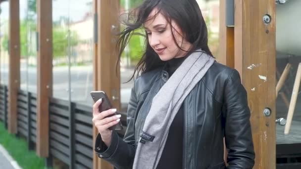 穿着时尚灰色围巾和黑色库尔斯克的漂亮年轻女孩看着手机 用手指按下按钮 长发在大风中发展 背景中的城市建筑 — 图库视频影像