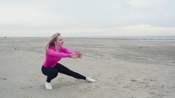 穿着黑色紧身裤的运动的漂亮女孩在荒凉的沙滩上伸展双腿的肌肉 在阴天炎热的夏日进行重训练前的体操 — 图库视频影像