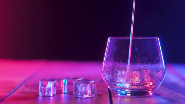 v průhledném skleněném skle pohár s kostkami křišťálového ledu, stojící na dřevěné stojan, je tenký potok nalit silnou voňavá whisky. spousta šplouchání ve skle. zavření, barevné osvětlení v liště.