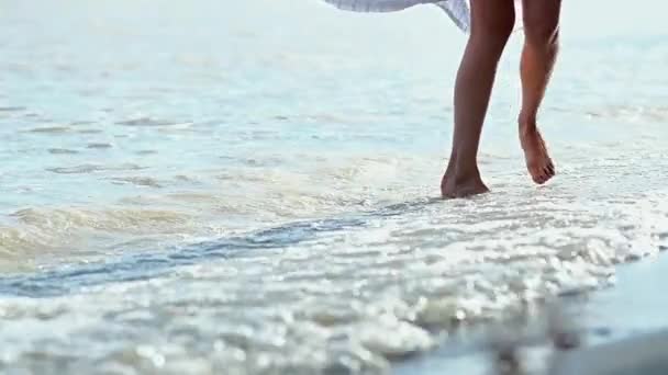 素足と白いドレスを持つ若いスレンダーな女の子は 海の砂浜に沿って歩きます サーフは女性のシンを打つと飛び散る 日没時のビーチで夏の日にスローモーション — ストック動画