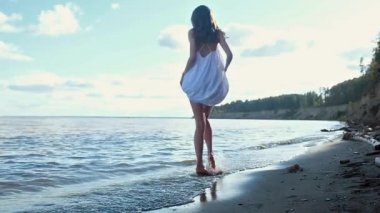 uzun bacaklar deniz kumlu plajda yürüyüş ile genç seksi model. Dönüyor, elleriyle beyaz bir elbise tutuyor ve gülüyor. Sörfü tekmeler ve suya su sıçratıyor. Denizde güneşli bir gün.