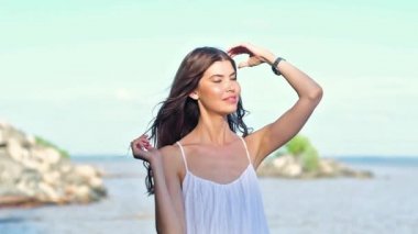 Anlamlı gözleri ile beyaz elbiseli Avrupalı genç kız, kameraya bakar, güler ve başını büker. Saçları rüzgara dolanmış. saçını düzeltir. Sahilde Sıcak Güneşli yaz günü.