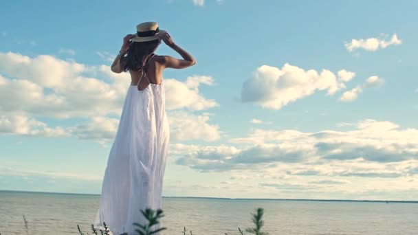 一个穿着白色衣服的漂亮苗条女孩脱下草帽 双手伸向两侧 摇摇长发 把脸转向太阳 背面的视图 温暖的海上日落 — 图库视频影像