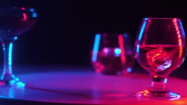 それらにアルコール飲料コニャック ウイスキー カクテル 冷たい立方体透明な氷が回転木製のスタンドに回転して それらに注がれた異なる形状のガラスタンブラー 多色照明 クローズアップ — ストック動画