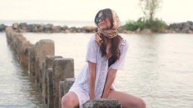 beyaz düğmesiz elbise ve siyah bir mayo genç güzel seksi kız, kafasına mendil ile, büyük siyah güneş gözlüğü elleri ile eğilerek dikdörtgen taşlar üzerinde oturur. Kameraya poz vermek
