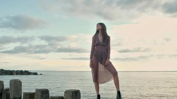 一个穿着透明棕色连衣裙的年轻美女 黑色的鞋子站在石柱上 在日落时分伸出水面 她拉直头发 看着相机性感 模特的晚间拍摄 — 图库视频影像