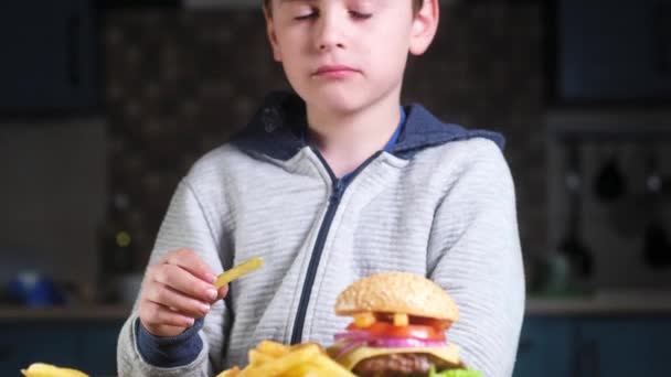 灰色のジャケットを着た少年がフライドポテトを食べている 木製のテーブルの隣には カツレツとチーズとケチャップのソーサーとジューシーなハンバーガーがあります — ストック動画