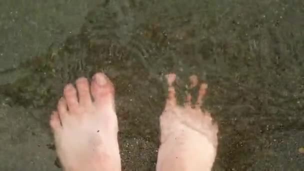 水の中で晴れた日にビーチに立っている裸足のヨーロッパの男性 透明な波が飛び散り 男は暗い砂の中で指を振る 上から見る — ストック動画