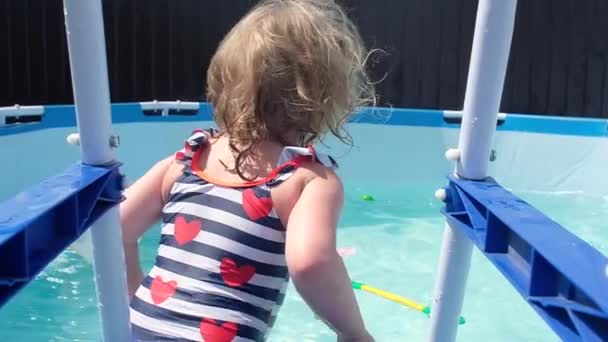 赤いハートの水着で軽い濡れた髪を持つ小さな女の子は カメラを見て プールの階段から膨脹可能な円でジャンプし 積極的に泳ぎ始めます 暑い夏の日に家の池で泳ぐ — ストック動画
