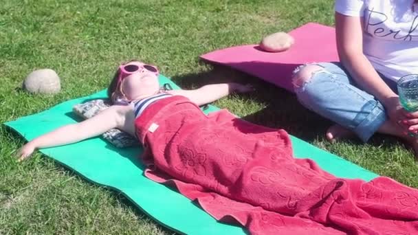 一个金发湿润的漂亮女孩 条纹泳衣 粉红色的太阳镜 躺在草地上 在妈妈旁边的一条红色的大毛巾下 笑着晒日光浴 大自然中的夏日休息日 — 图库视频影像