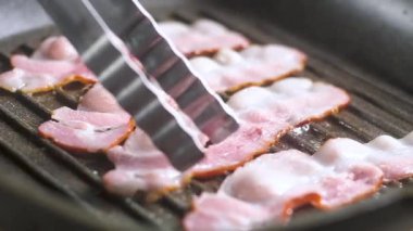 Şef adam metal mutfak maşaları düzelten yağ sulu domuz pastırması lezzetli ince kokulu dilimleri, hangi yapışmaz kaplama ile demir bir tavada ızgara üzerinde ısıtılır. Jambon dan buhar ve duman gider.