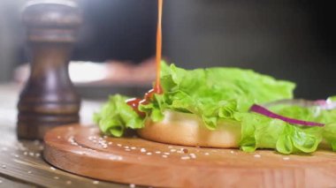 şef ahşap standı üzerinde yatan topuz ile taze salata yeşil yaprak üzerine domates sosu döker. sonraki karabiber biber shaker olduğunu. mutfakta Burger Pişirme süreci. Yavaş çekim yakın çekim. Yapay aydınlatma.
