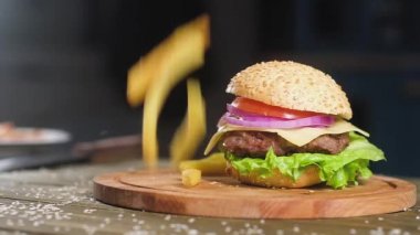 Kızarmış uzun Patates kızartması salata, peynir, gevrek pastırma ve ahşap bir tepsi üzerinde yatan kızarmış pirinci ile pişmiş Burger düşmek. Mutfağın arka planında yavaş hareket.