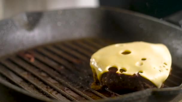 シェフのスパチュラは 沸騰油のスプレーでノンスティックコーティングとホットフライパングリルとサクサク茶色の地殻とジューシーな肉のパテのペアをピックアップします 穴が開いている黄色のチーズの薄いスライスの上に — ストック動画