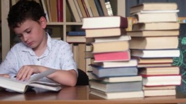 koyu saçlı çocuk, açık gömlek masada oturur ve kitap okur. Özlü -yor. Yanak üzerinde el güvenerek sayfaları ile Yapraklama. kitap yığını nda bir sürü kitap. Kitaplığın arka planında panoramik çekim.