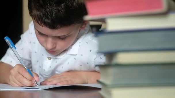 坐在木桌前的男孩的特写镜头 他在笔记本上用一支蓝色的钢笔写字 解决教室里的问题 一堆书里有很多课本 学习过程全景拍摄 — 图库视频影像