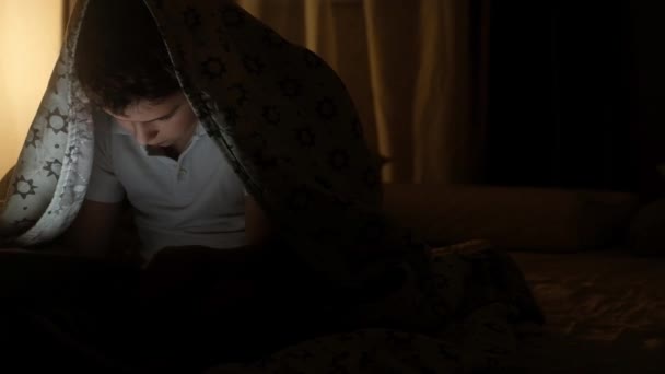 dítě v noci v bílém tričku, které sedí na posteli pokryté přikrývkou, kouká na tabletu a směje se. V pozadí svítí noční světlo. Panorama průměrného plánu.