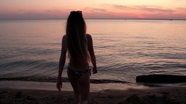 長い髪が砂の上を歩いている色の水着を着た若いスレンダー美少女 海に来て 両手を横に上げる 砂浜の美しいカラフルな夕日の背景 バックライト — ストック動画