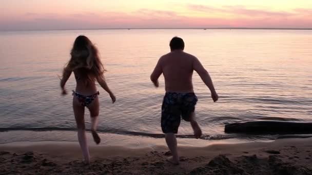 ショートパンツの太った男と水着の楽しい若いスレンダーな女の子が海に走ります 砂浜のカラフルな夕日 バックライト — ストック動画