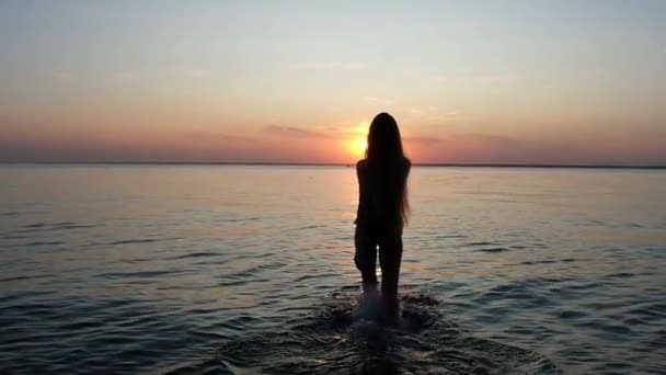水着姿のスレンダーな女の子のシルエットと長い髪で 美しい夕日に対してゆっくりと海に入ってくる — ストック動画